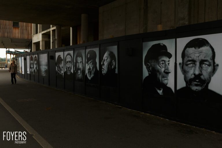A long black wall displays a series of black and white portrait photographs of men's faces in an urban underpass setting.