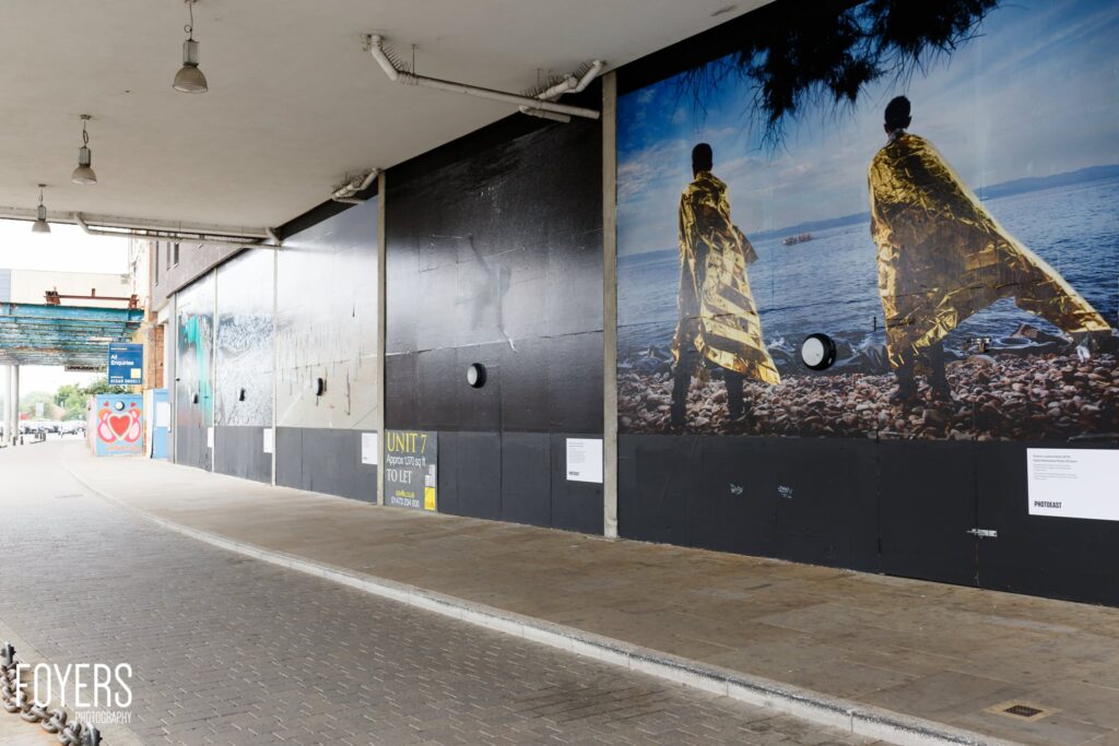 Large outdoor mural on a warehouse wall showing two figures in gold foil blankets standing on a pebbly beach facing the sea.