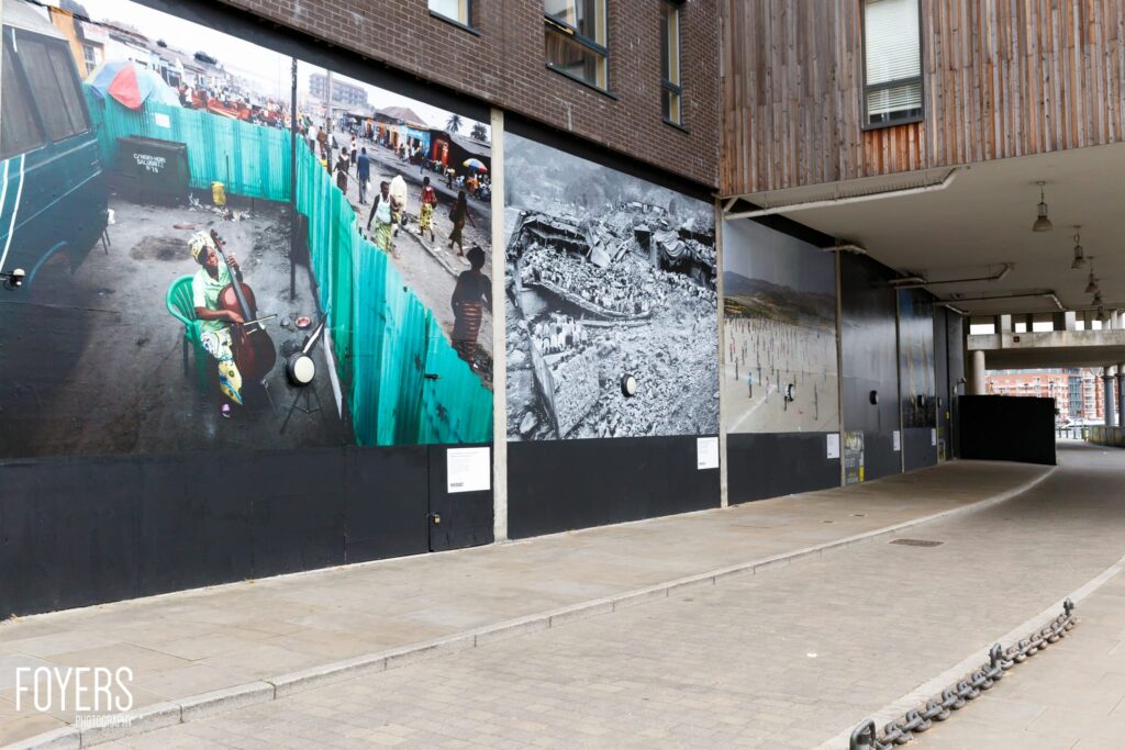 Large-scale photographs displayed on a dark hoarding in an urban courtyard between brick buildings.