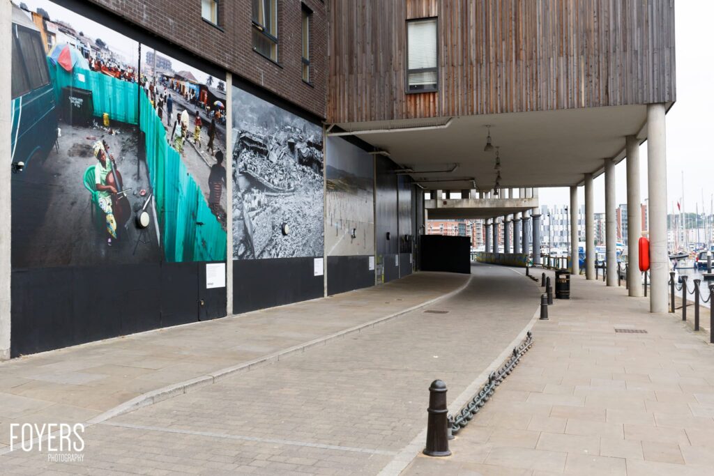 Underpass with large photographic displays on wall, concrete pillars, and marina visible in background.
