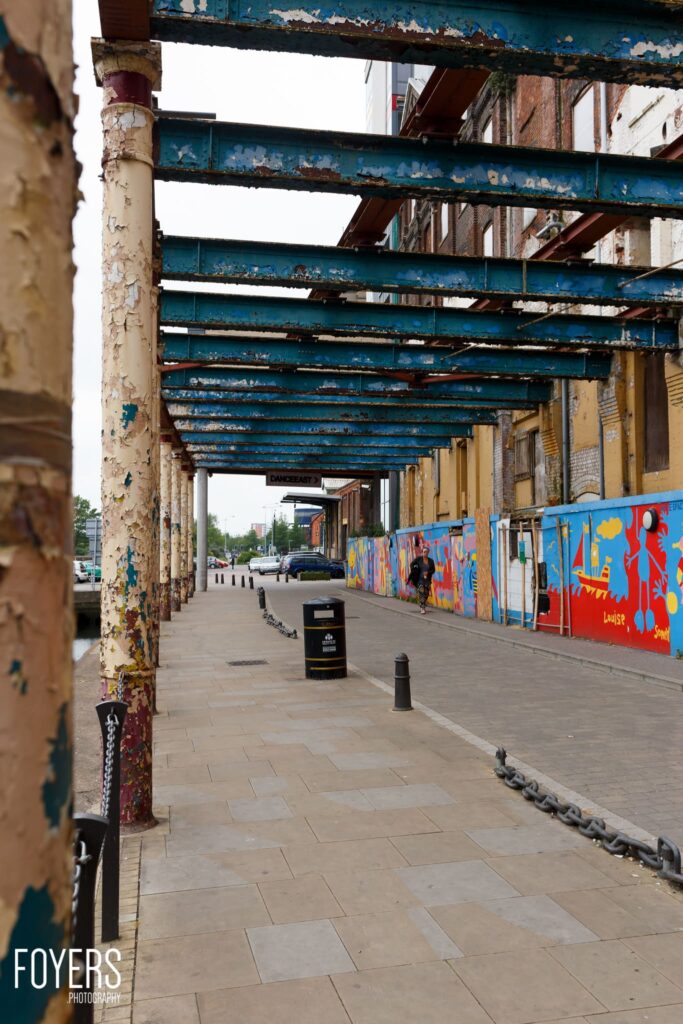 An underpass with peeling blue steel beams, rusty pillars, and colorful street art on walls. A few pedestrians and vehicles visible in the distance.
