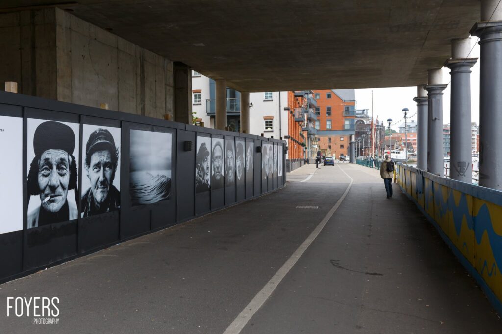 Underpass with black and white portrait photographs displayed on panels along the left wall, urban waterfront visible ahead.