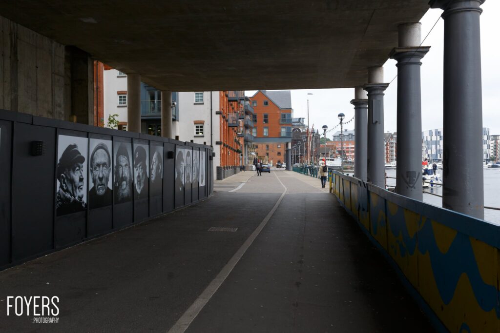 A pedestrian bridge beneath a concrete overpass features black and white portrait panels on the left wall and colorful chevron markings on the right railing.