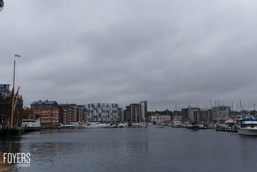 A marina with moored sailboats and yachts, waterfront buildings, and overcast sky. Photographer credit: Foyers.