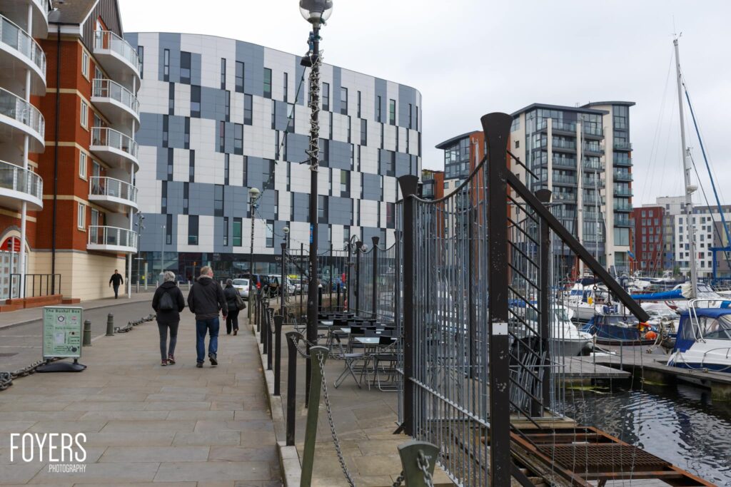A waterfront promenade with modern apartment buildings, moored boats, and a few pedestrians walking along the quay on an overcast day.