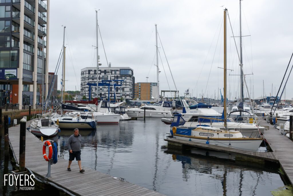 A marina with moored sailing boats, a person jogging on the wooden dock, modern apartment buildings in the background under an overcast sky.