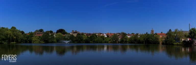 A riverside town with brick buildings and a church spire reflected in calm blue water, under clear sky.