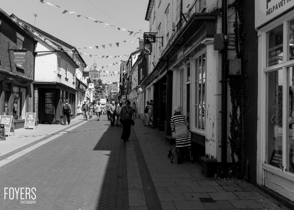 A busy pedestrian shopping street in black and white, lined with historic buildings and bunting overhead.