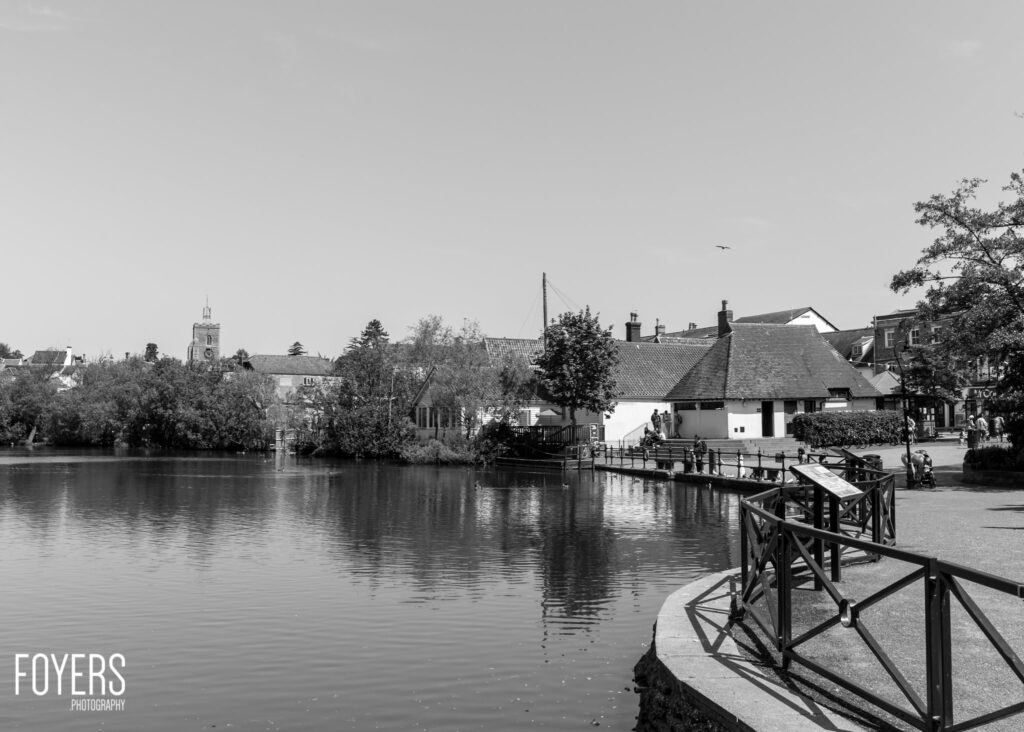 A riverside town scene with wooden railings in foreground, calm water, historic buildings and church tower across the river.