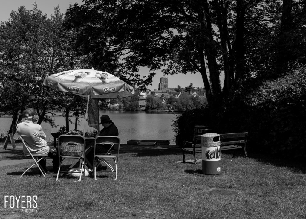 People picnicking by a river under a Kelly's branded umbrella, with a bin and benches nearby, trees framing the scene.