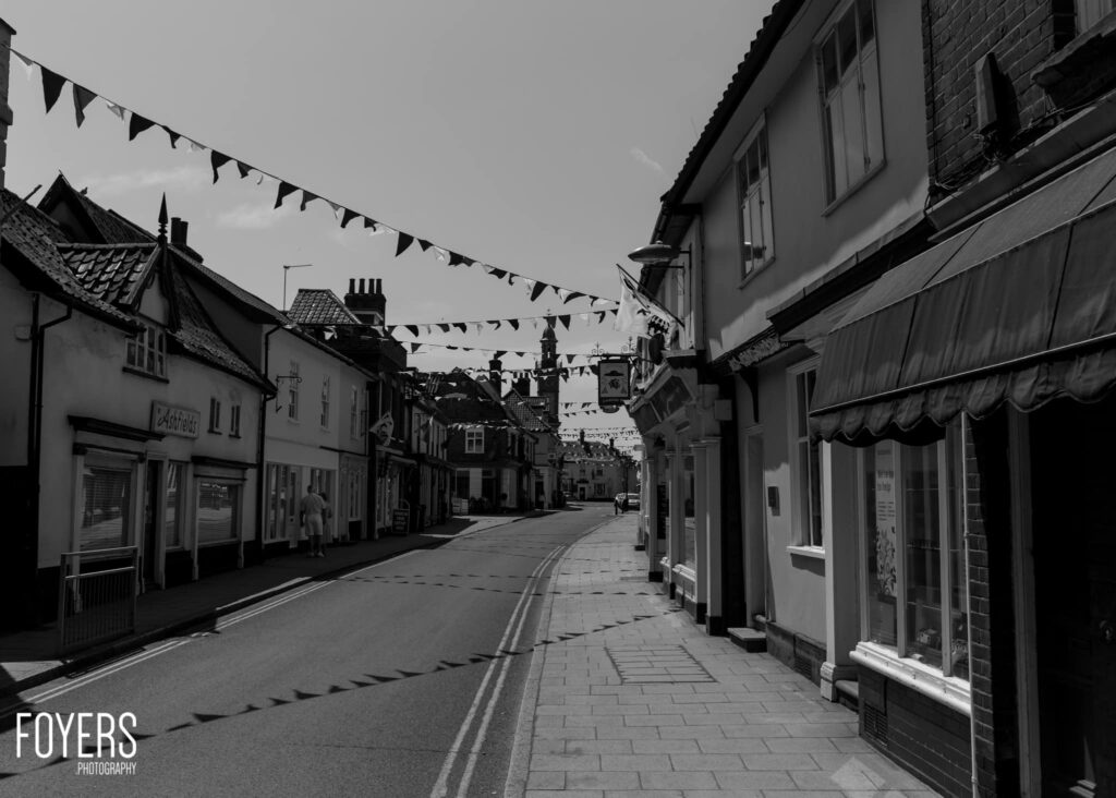 A quiet British high street with shop fronts on both sides, bunting overhead, and a church tower visible in the distance.