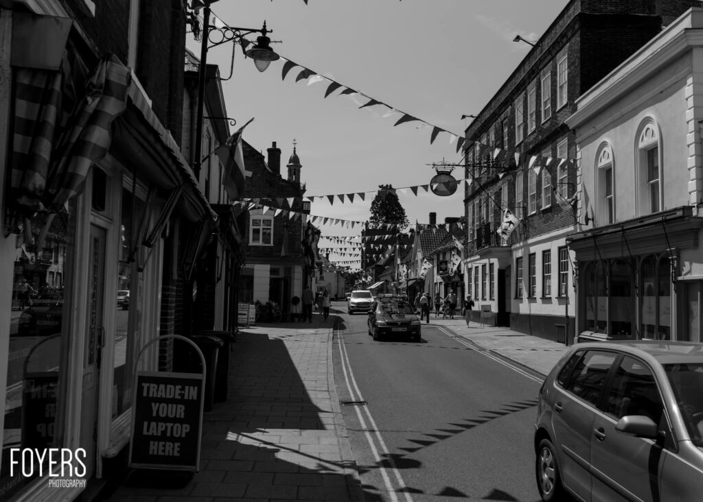 A high street lined with traditional buildings decorated with bunting. Cars parked along the road, shops visible on both sides.