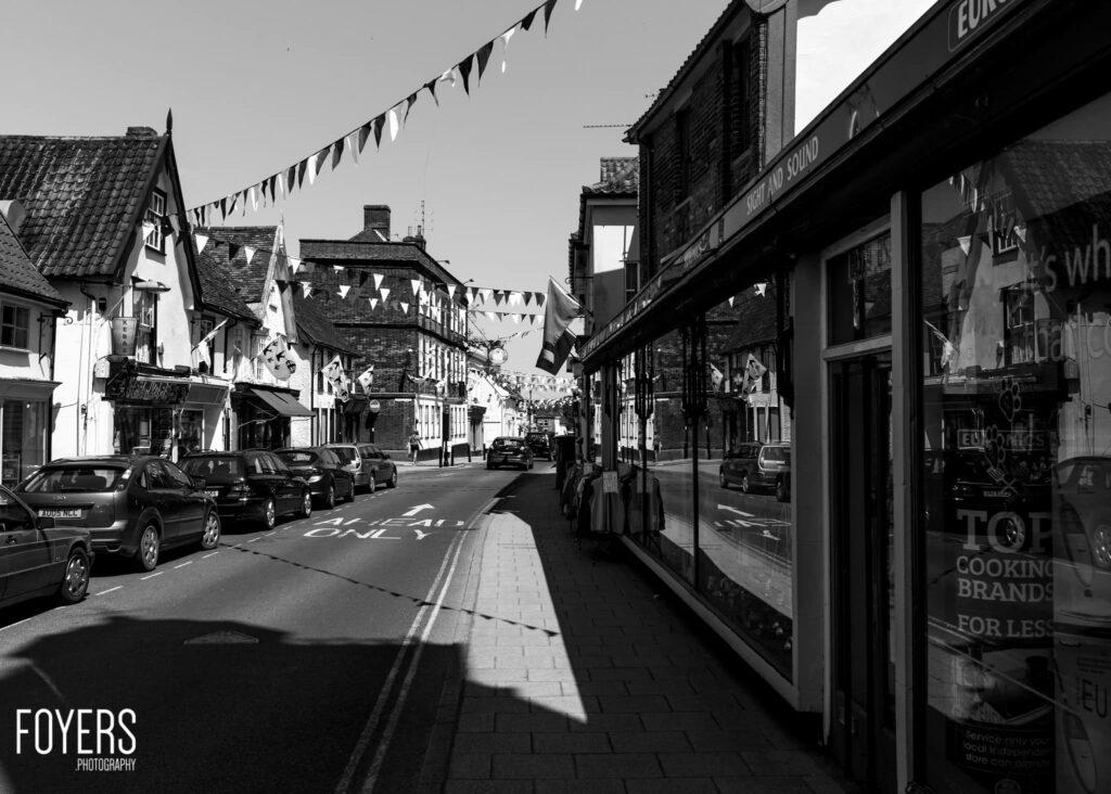 A black and white street scene showing a historic high street with period buildings, parked cars, bunting overhead, and shop fronts on a sunny day.