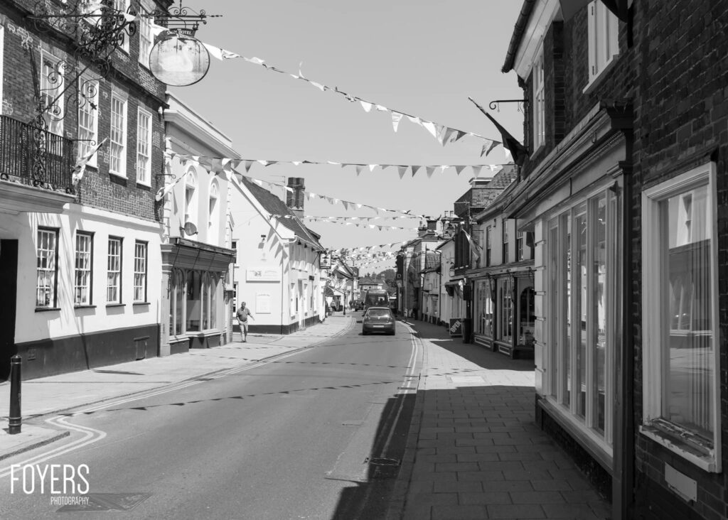 A quiet high street lined with brick buildings and shop fronts, decorated with bunting overhead, a car parked mid-street.