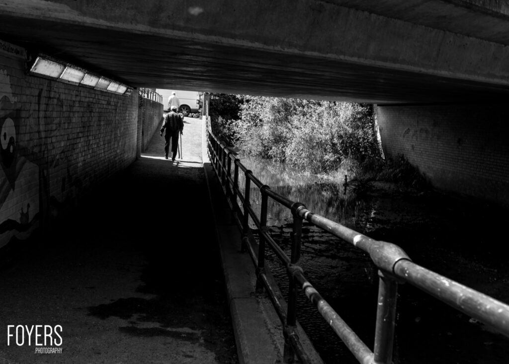 A person walks through a dark underpass towards bright daylight, with metal railings and brick walls visible.