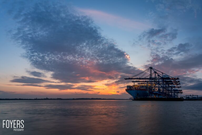 A large container ship at dock during sunset, with dramatic orange and blue clouds reflected in calm water.