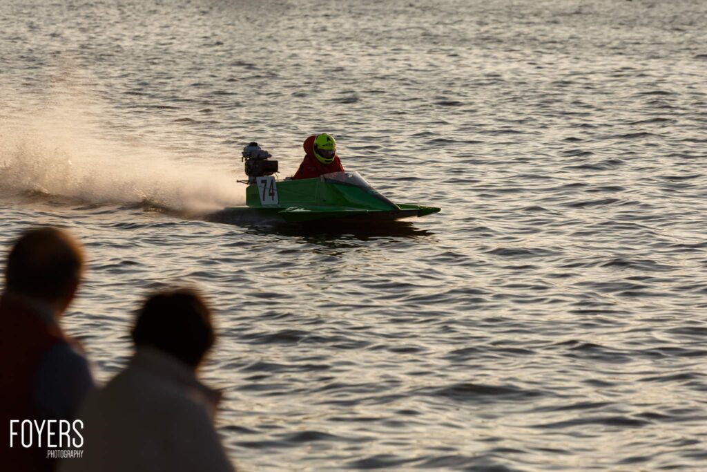 A jet ski racer in a green and black vessel numbered 74 speeds through ocean waves, observed by spectators on shore.