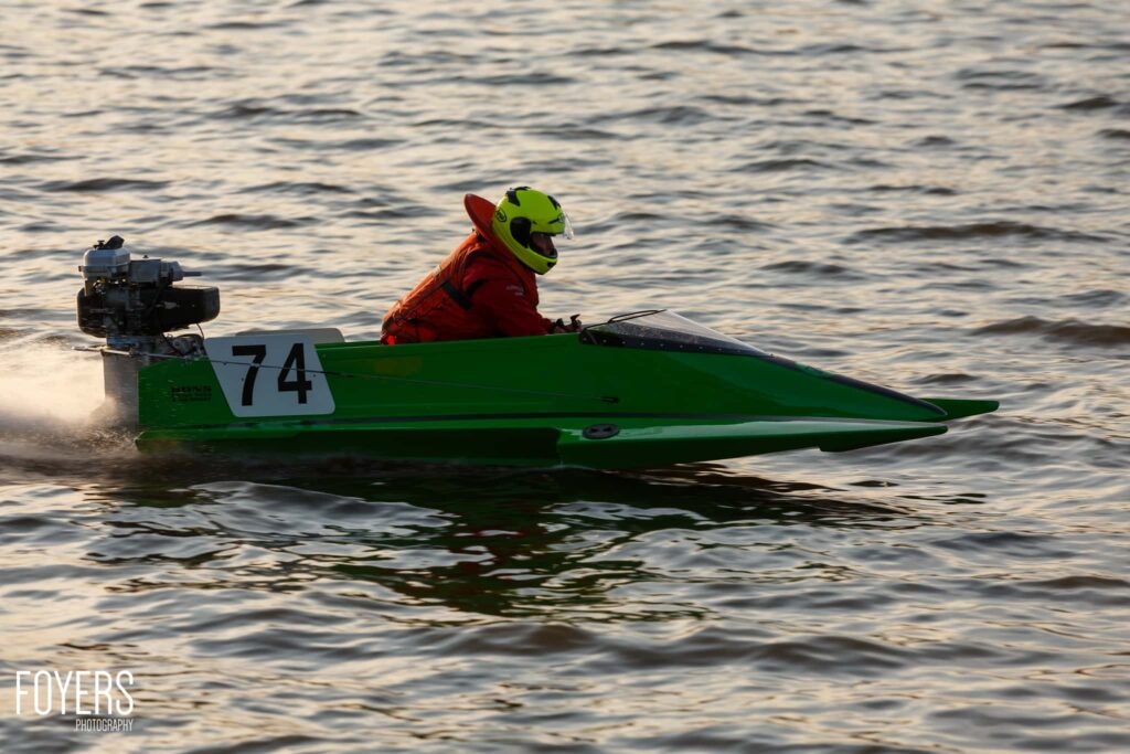 A green speedboat numbered 74 with a driver in a lime helmet and red jacket racing through shallow water.