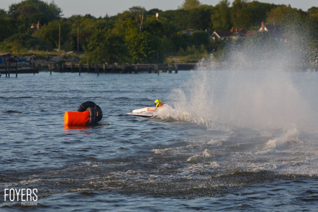 Jet skier riding a wave near an orange buoy, with a moored boat and riverbank visible behind.