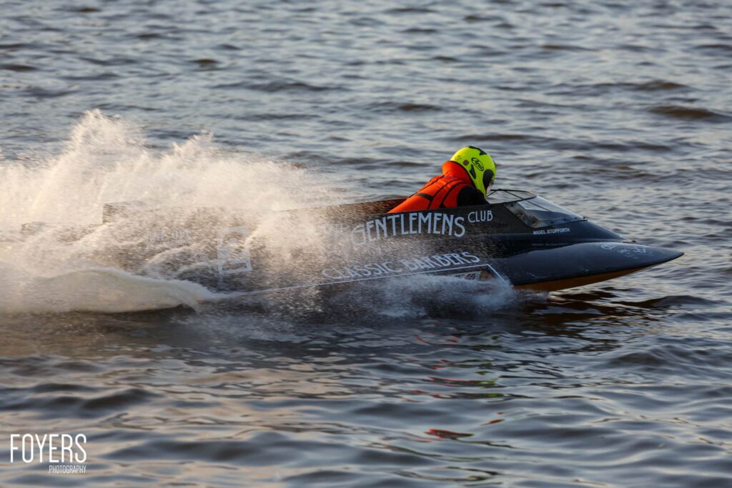 A jet skier in a neon yellow helmet and orange life jacket rides through a wave on a black jet ski branded with Gentlemens Club.