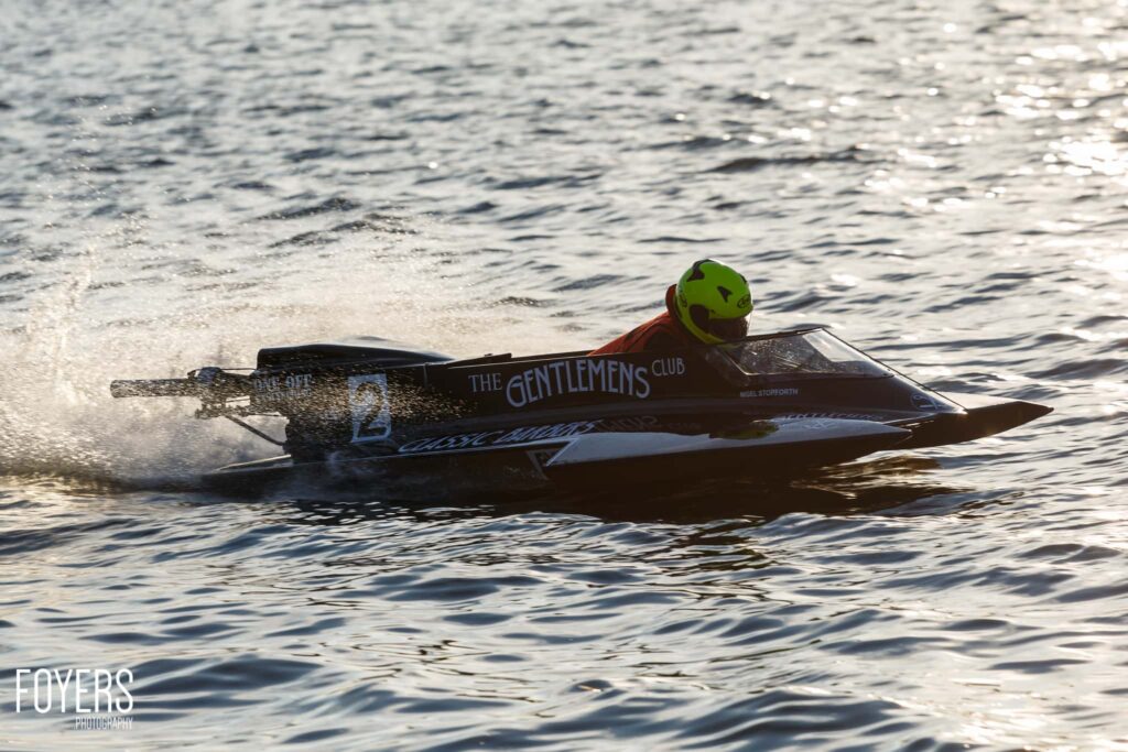 A speedboat named The Gentlemens Club races through shallow water with a helmeted driver aboard, creating spray.
