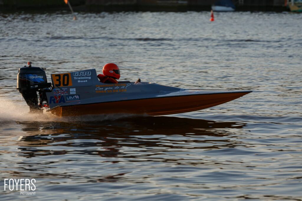 A speedboat with number 30 and Training Boat written on its side speeds through shallow water near a beach, with one person aboard wearing a red helmet.