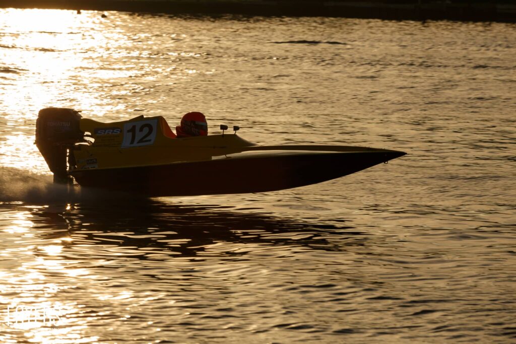 Racing speedboat numbered 12 silhouetted against golden water during sunset or sunrise.