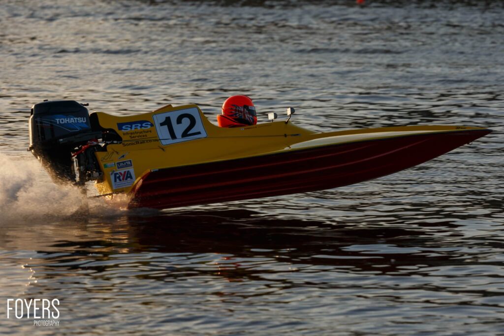 A yellow and red speedboat with race number 12 speeds through shallow water, piloted by someone in a red helmet.