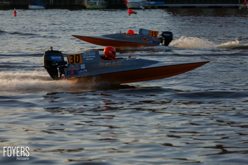 A speedboat with blue hull and number 50 approaches the beach in shallow water, pilot wearing red helmet visible.