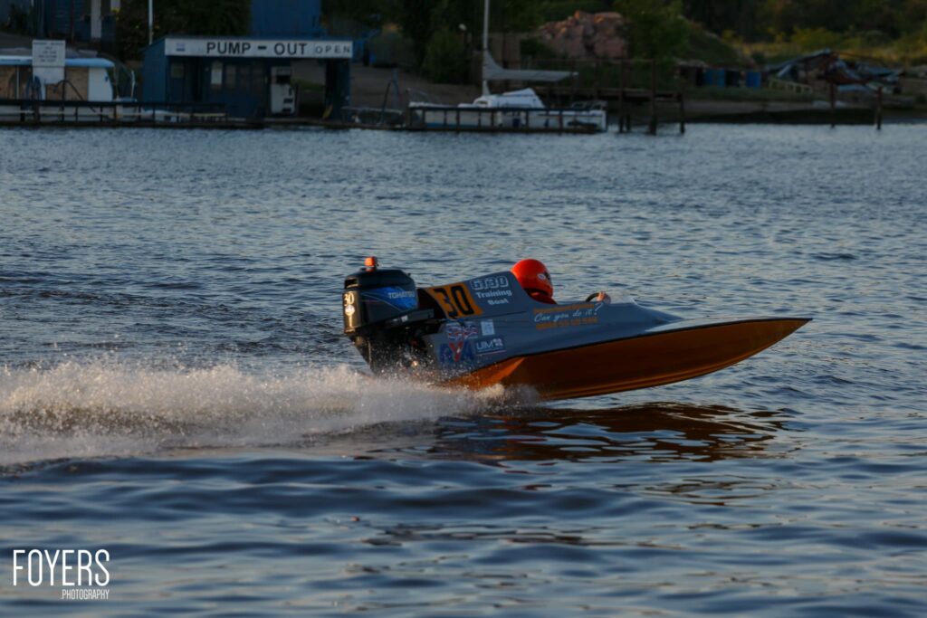 A speedboat with blue and orange hull approaches the shore, creating spray as it enters shallow water near a pump-out facility.