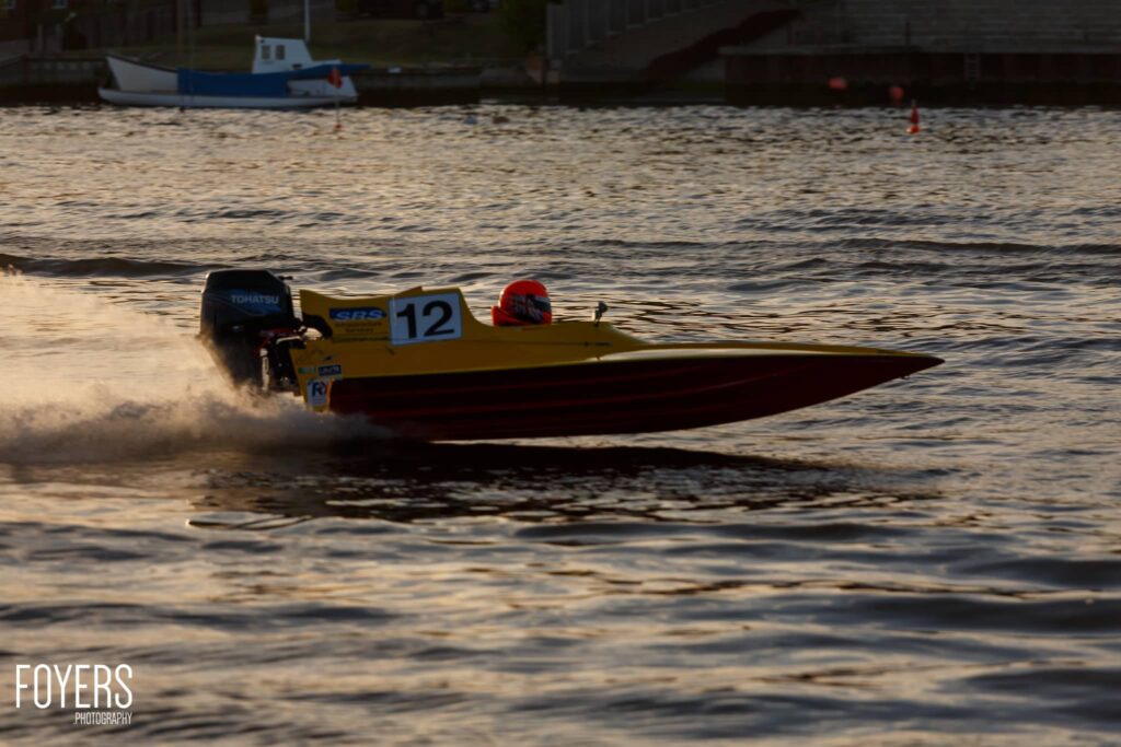 Racing speedboat numbered 12 with yellow hull and red trim speeds through shallow coastal water, creating spray.
