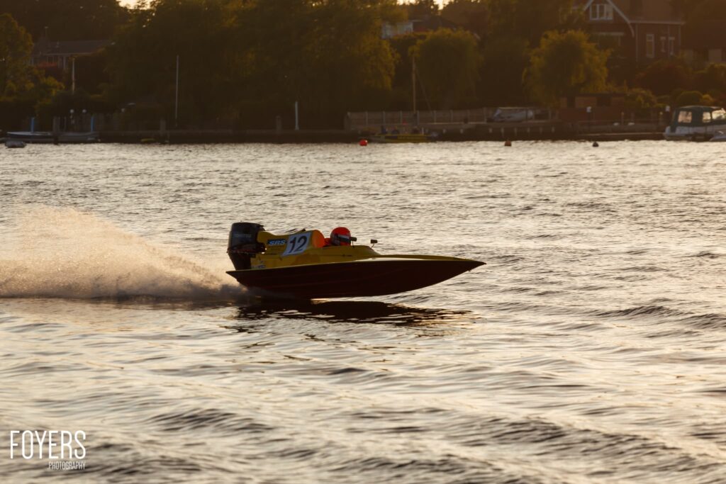 A yellow speedboat with number 12 races through shallow water near a beach, creating a large spray wake.