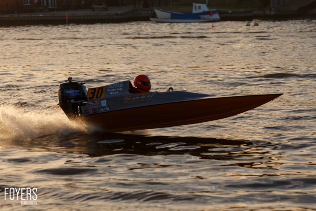 A speedboat with a red-helmeted driver races through shallow water near a beach, creating a spray of water.