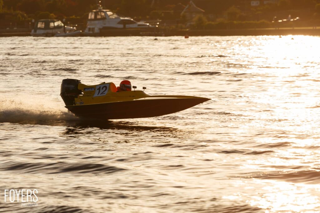 A yellow speedboat with number 12 races through shallow water near a beach at sunset, with moored boats visible in the background.