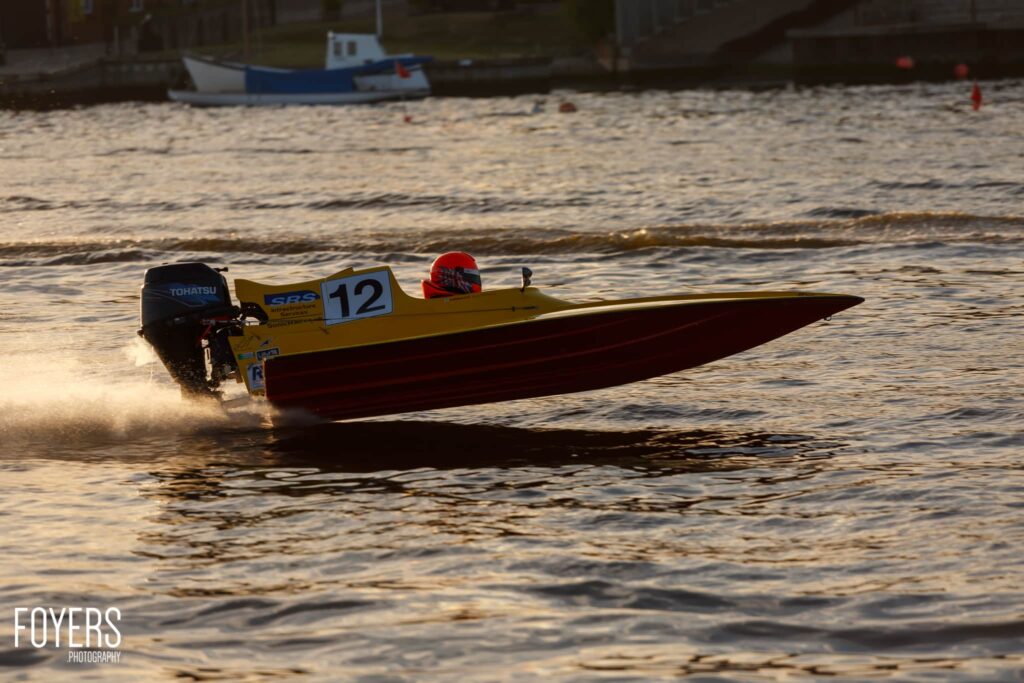 Racing speedboat numbered 12 with yellow hull and red trim speeds through shallow water near a beach, pilot wearing red helmet visible.