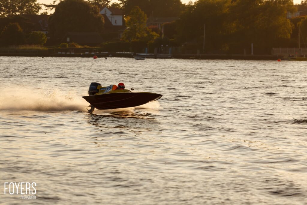 A speedboat with two occupants skims across choppy water near a riverbank lined with trees and buildings at dusk.