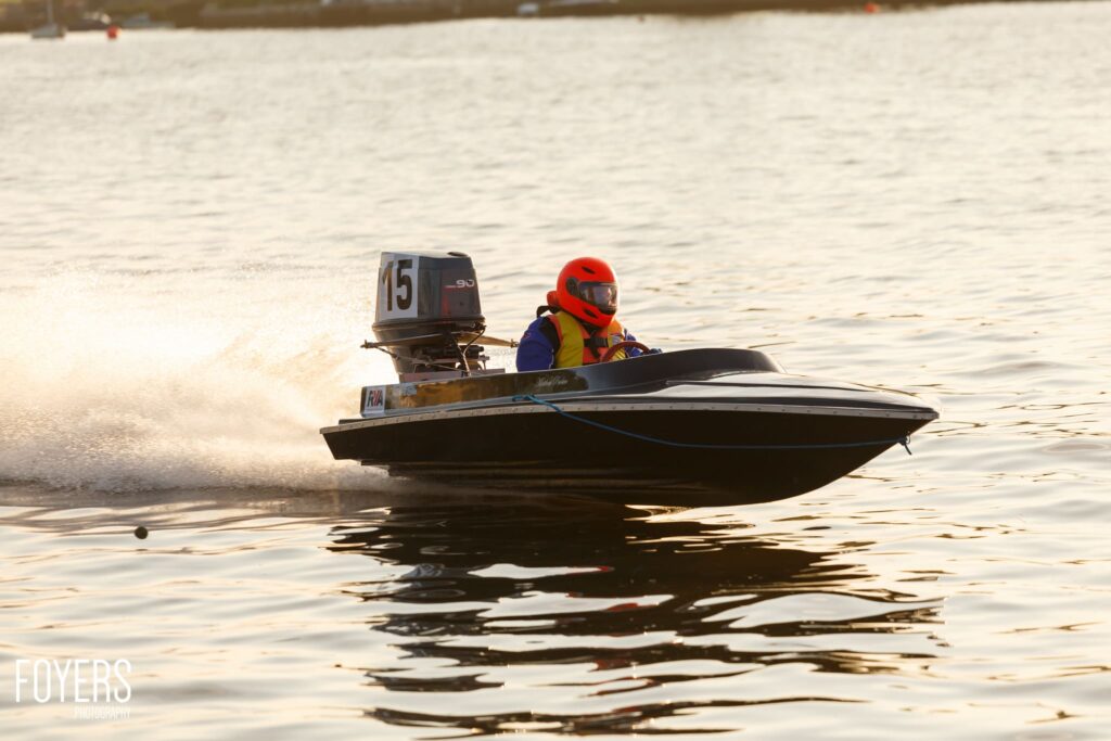 A speedboat with number 15 on its engine races across calm water, piloted by a person wearing a red helmet and life jacket.