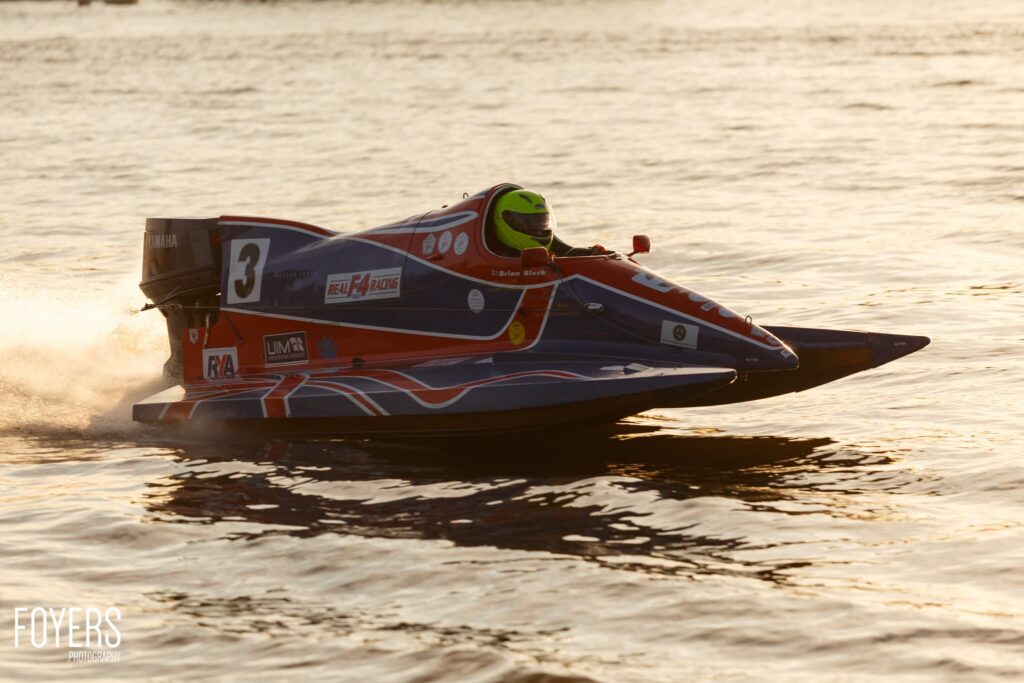 A red and black speedboat with number 3 races across shallow water, creating a spray of water behind it.
