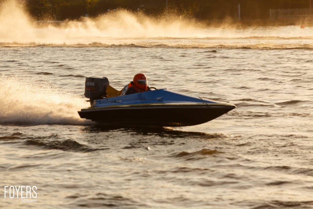 A speedboat with a blue hull and black trim speeds through shallow coastal waters, with a operator in an orange helmet and life jacket visible. Spray kicks up behind the boat as waves break in the background.