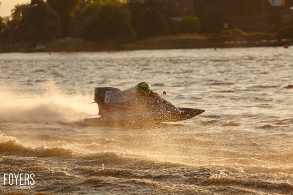 A jet ski rider in bright green goggles speeds through choppy brown water, creating a spray of water around them.