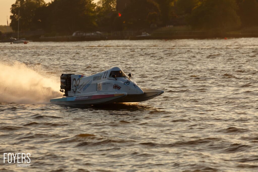 A speedboat with number 22 races across choppy water, creating a spray of white foam. Trees line the distant shore.