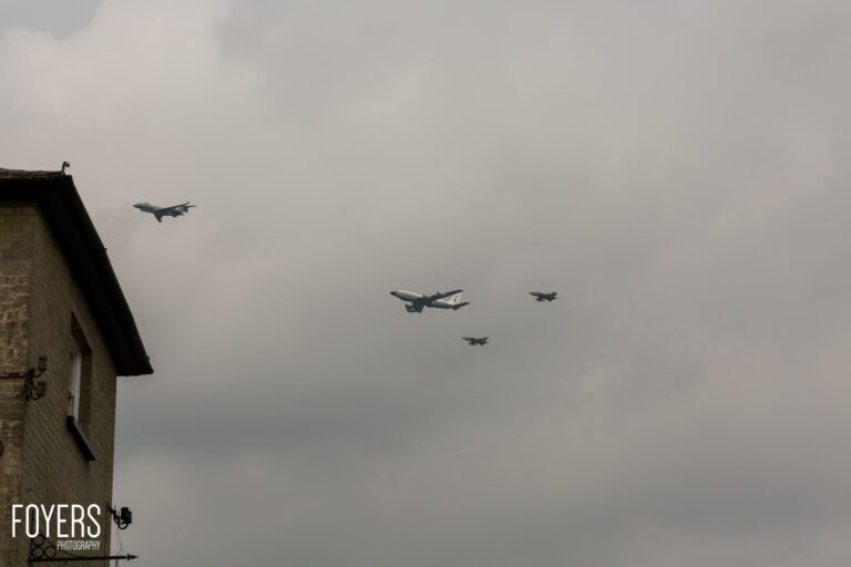 Military aircraft in formation flying over a brick building against a grey sky.