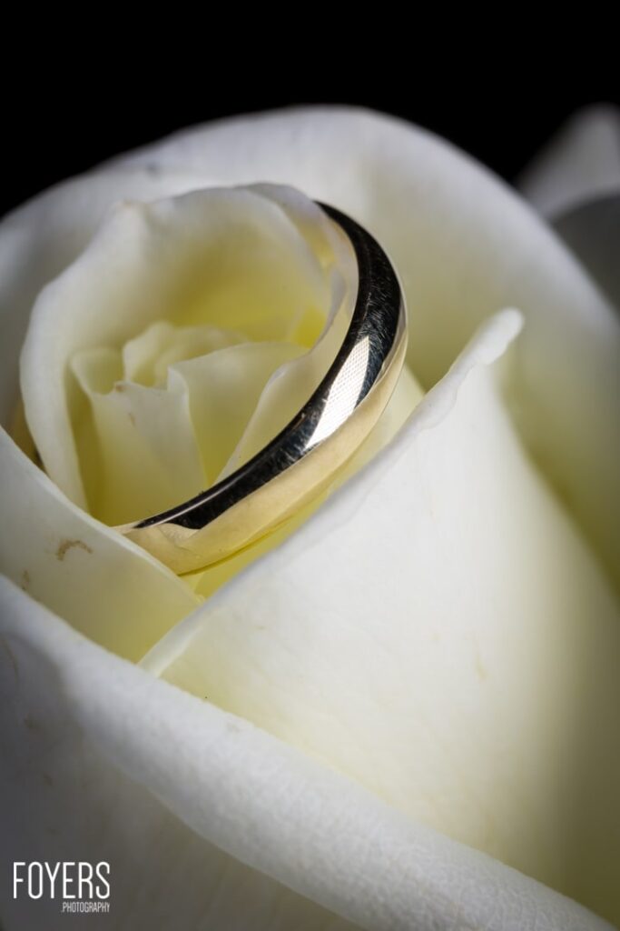 A wedding ring rests on a white rose petal, with soft petals blurred in the background.