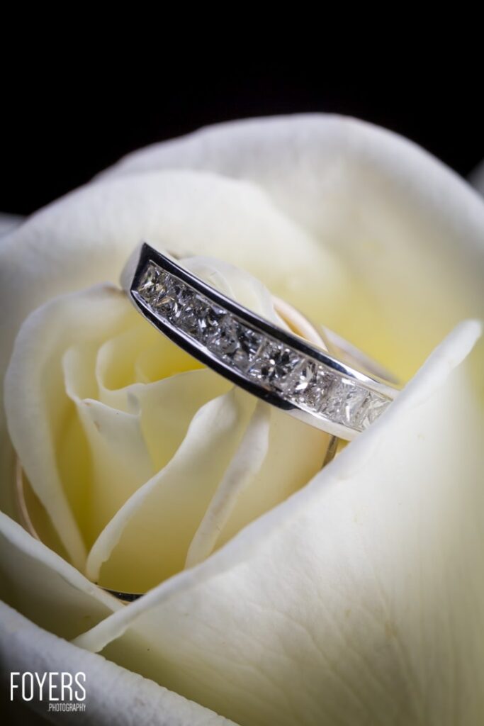 A diamond wedding ring rests on the petals of a white rose against a black background.