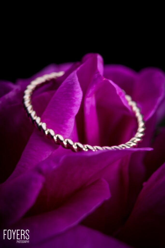 A vibrant magenta rose with delicate petals and a decorative pearl-edged ring wrapped around its bloom against a black background.