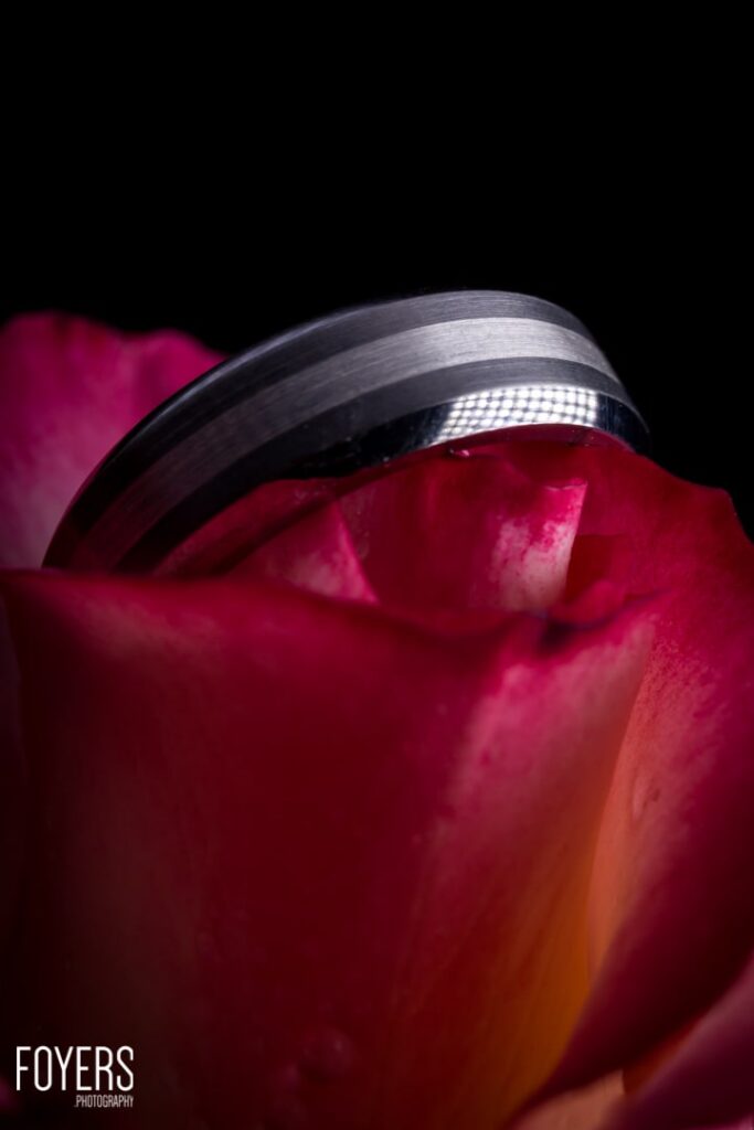 A dark metallic ring rests on deep red rose petals against a black background.
