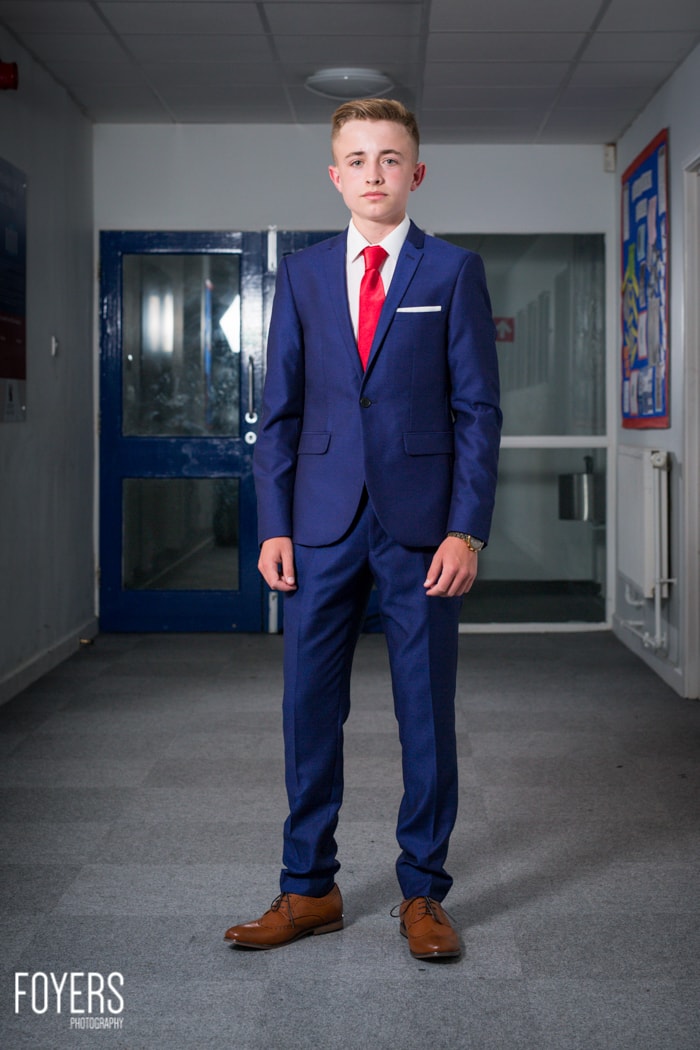 A young man in a navy suit and red tie stands in a school corridor, looking at the camera.