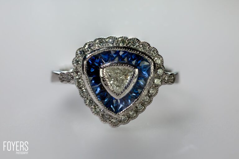 A heart-shaped diamond ring with sapphire surround and diamond halo, displayed on a light grey background.