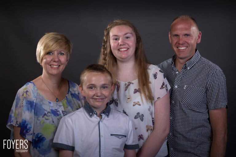 A family of four smiling at the camera against a dark background. Professional studio portrait photograph.