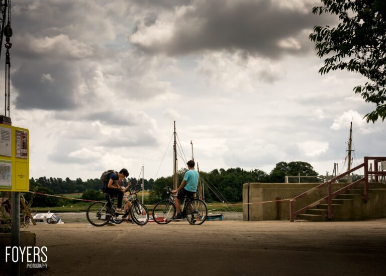 Two cyclists with bikes on a waterfront path, with moored sailing boats and a concrete bridge structure behind them.